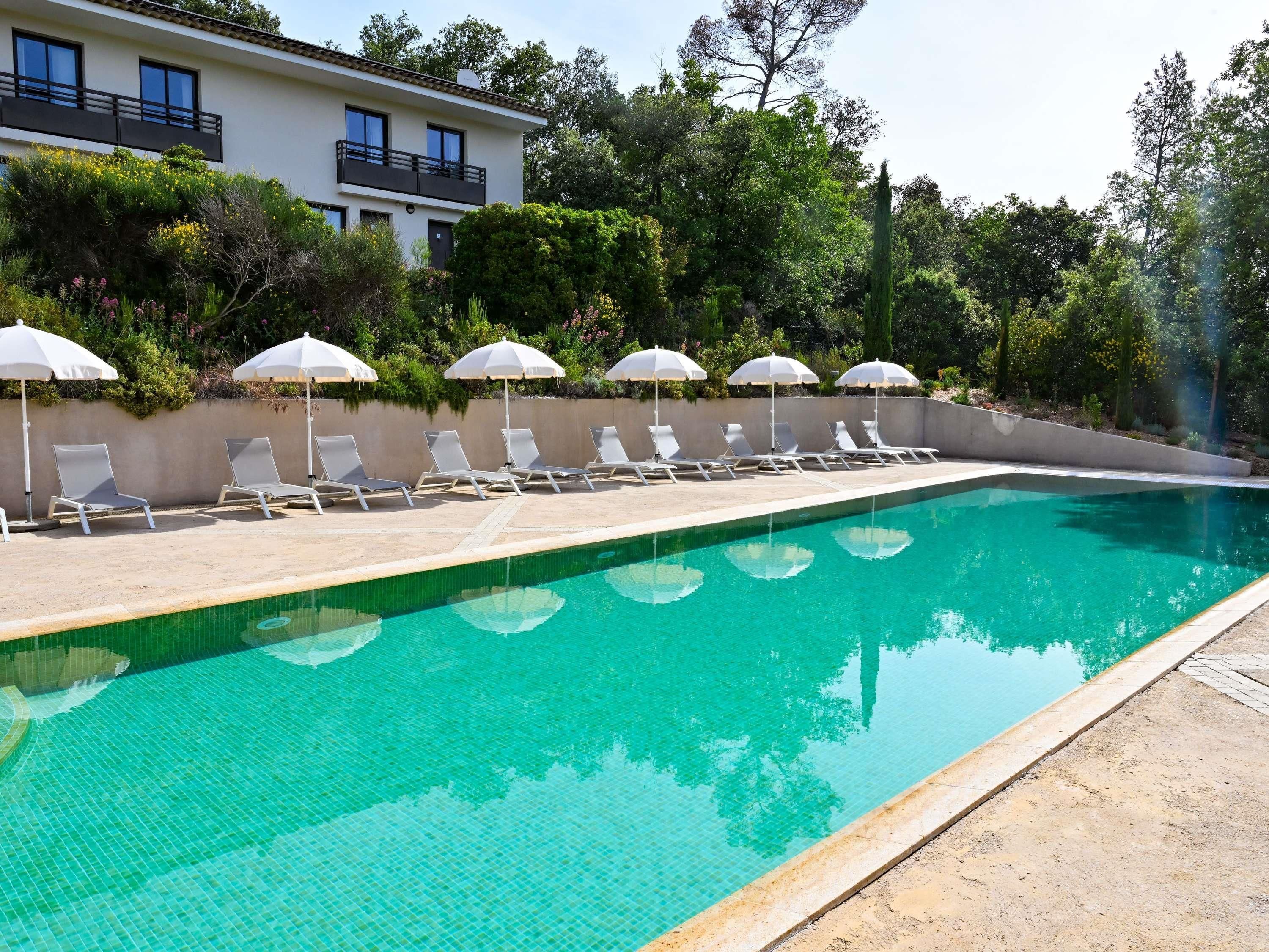 Una piscina con sillones de descanso y sombrillas, rodeada de vegetación y un edificio.