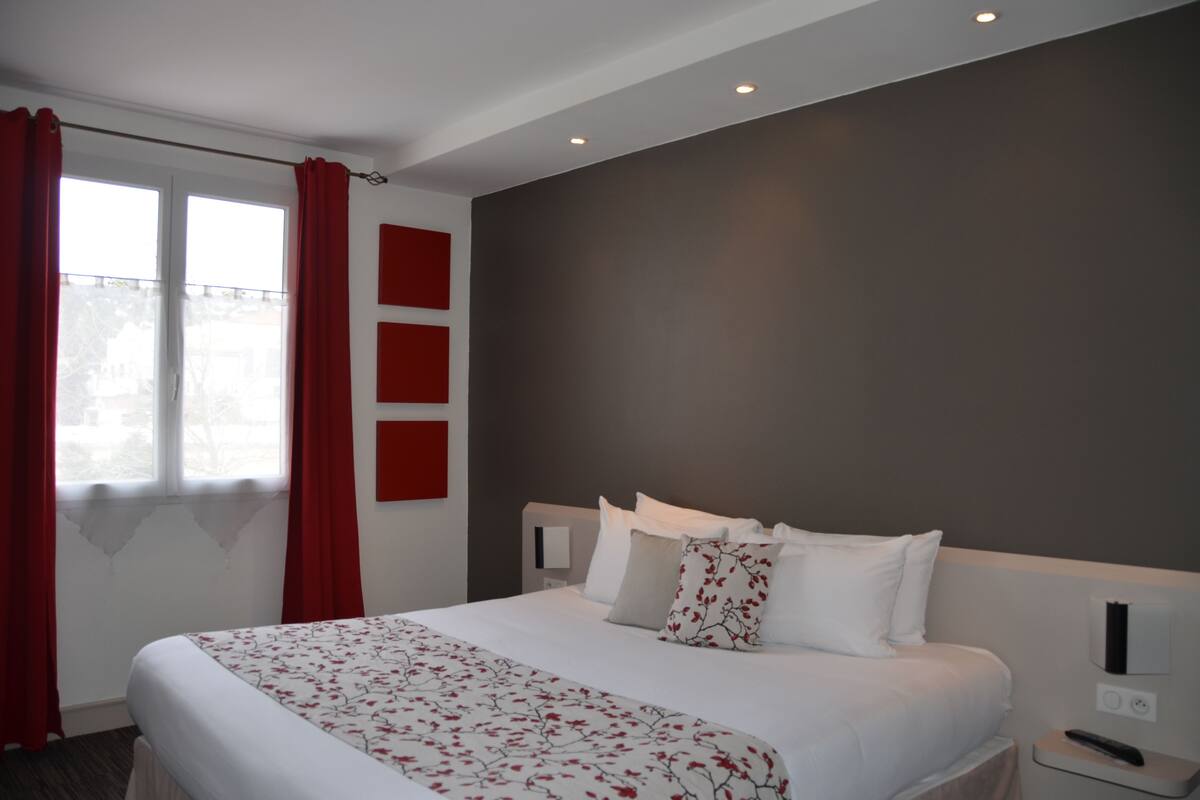 Ein modernes Hotelzimmer mit einem großen Bett, roten Vorhängen, einer grauen Akzentwand und einem kleinen Nachttisch mit Fernbedienung.