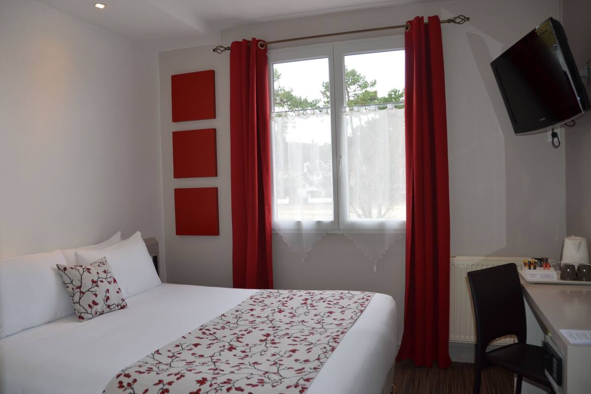Ein Hotelzimmer mit einem Bett, einem Schreibtisch mit Stuhl, einem Fernseher und einem Fenster mit roten Vorhängen.