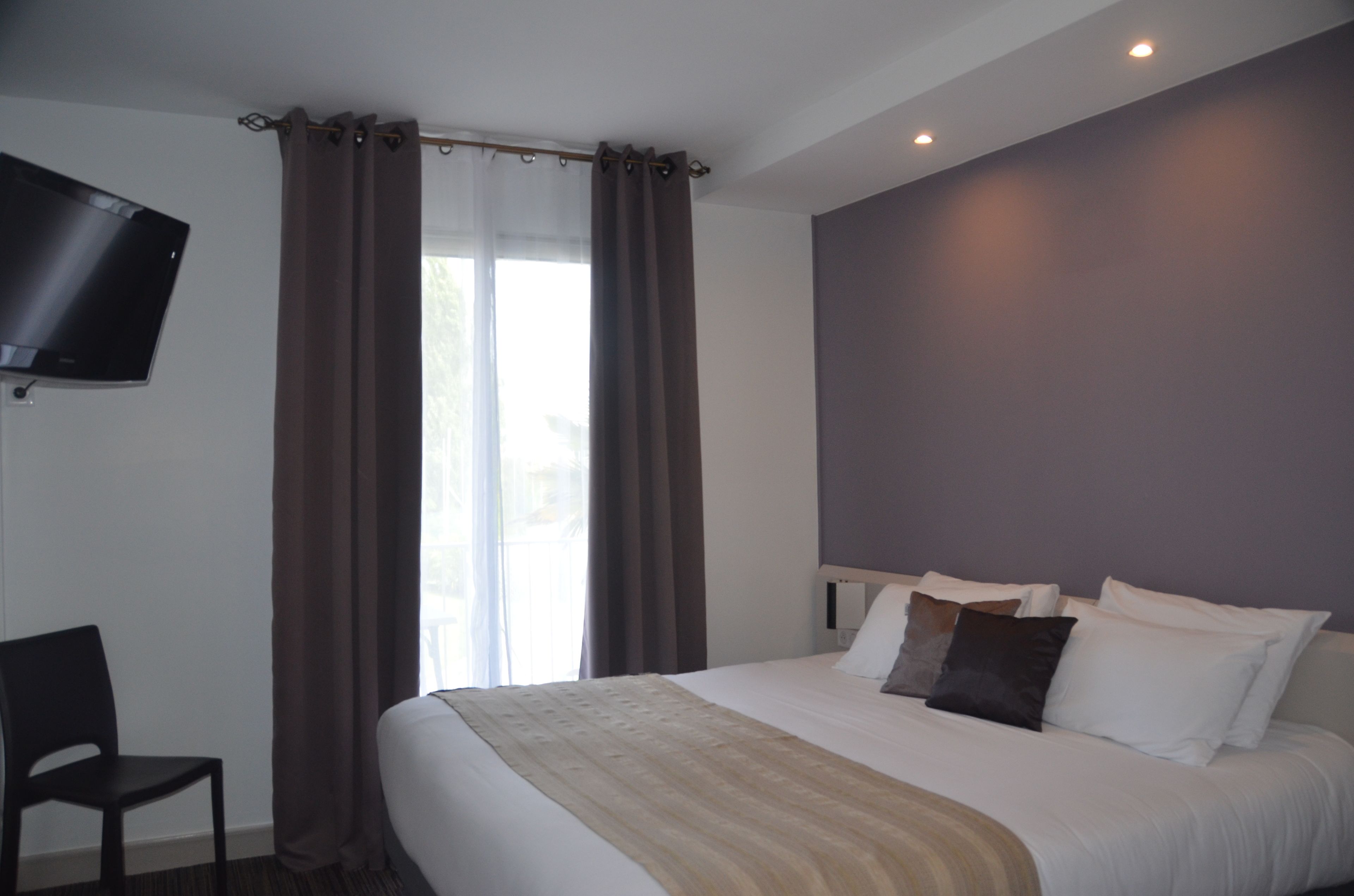 Habitación de hotel con cama, una silla, un televisor de pantalla plana en la pared y una ventana con cortinas.