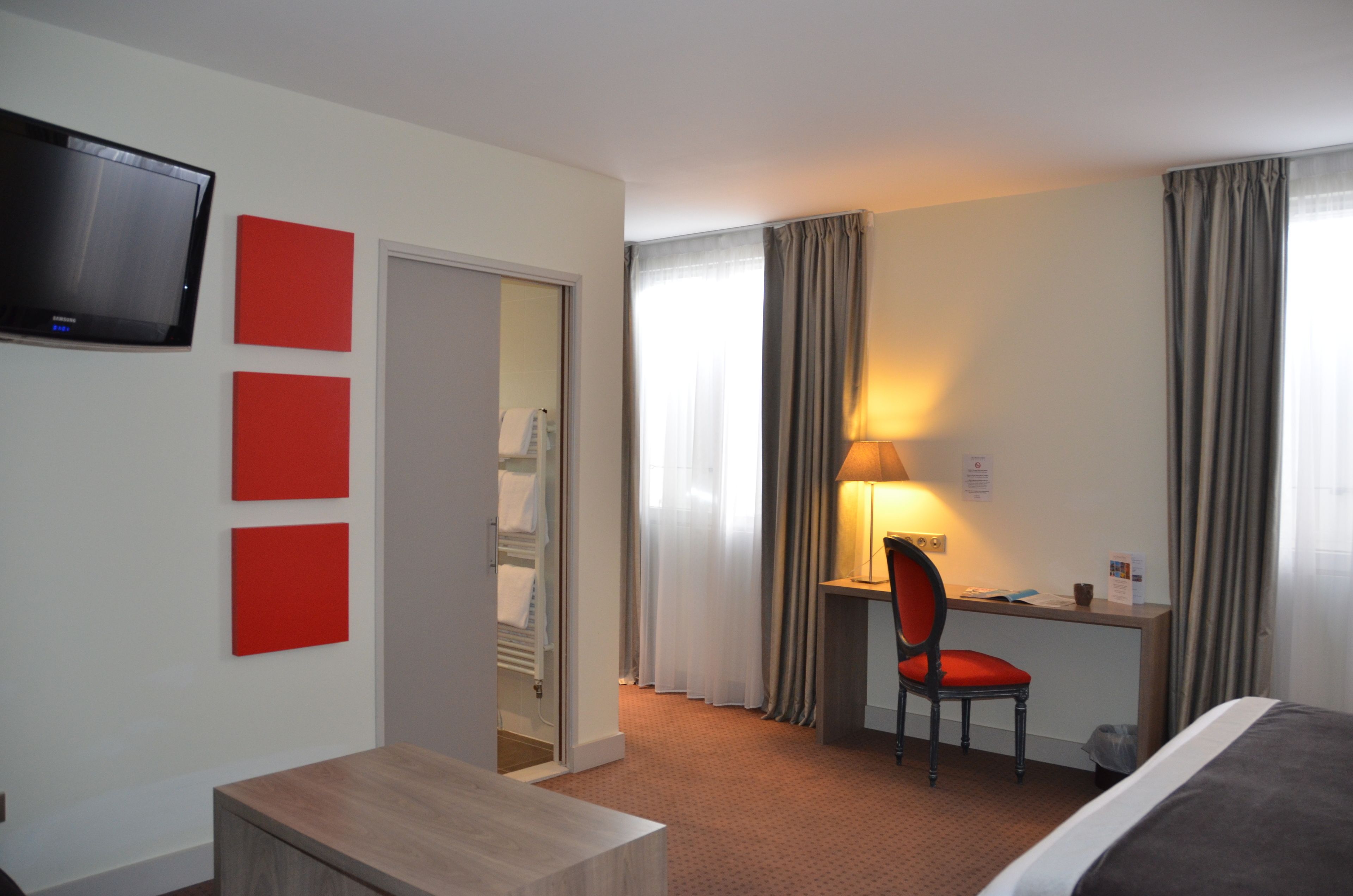 A hotel room with a bed, a desk with a chair, a television mounted on the wall, and a door leading to a bathroom.