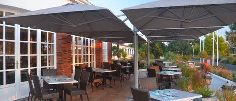 An outdoor patio with tables and chairs under umbrellas, surrounded by brick walls and glass doors.