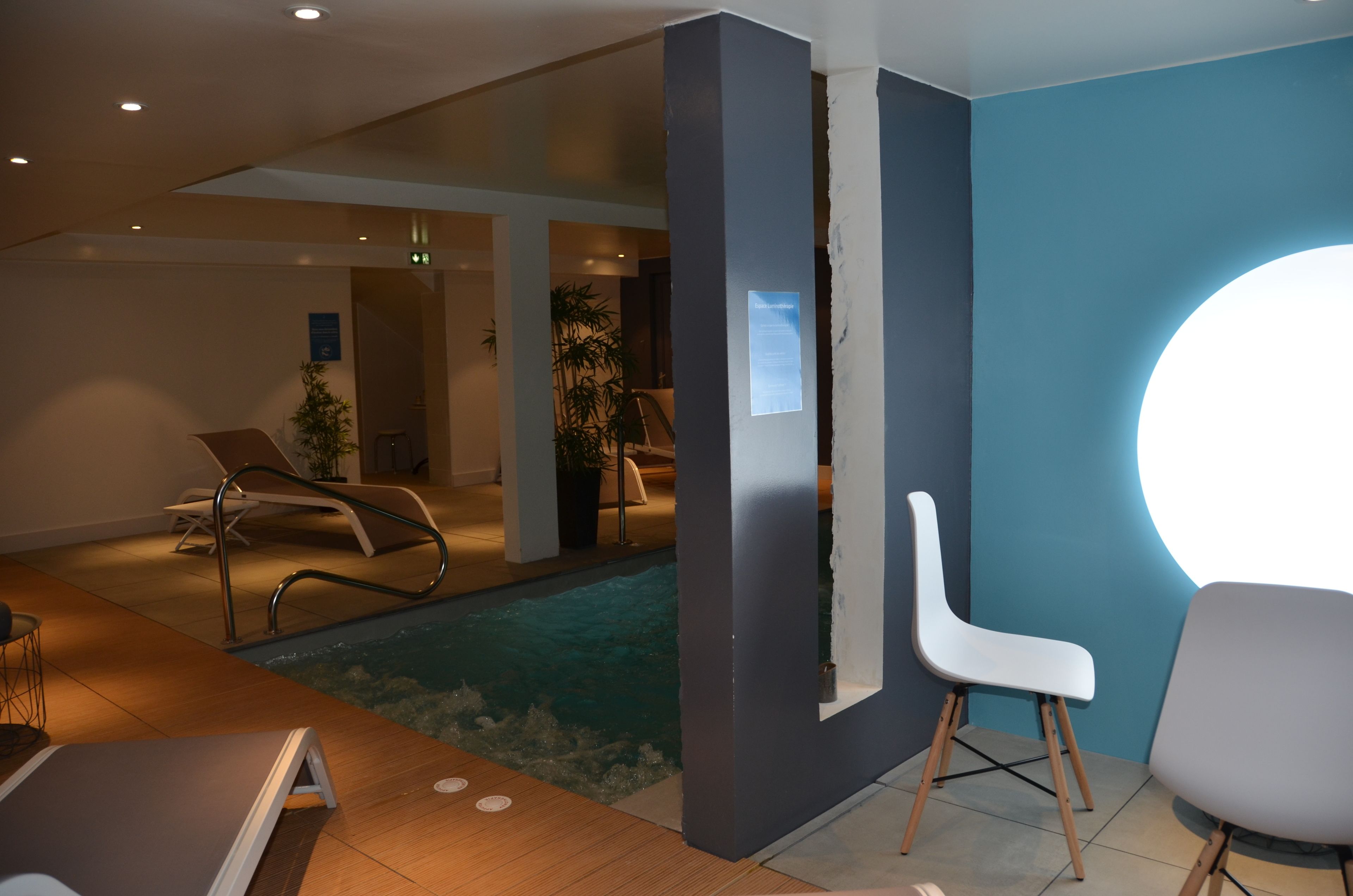 Un área de piscina interior moderna con sillones de descanso, suelo de madera y pared azul con un aplique circular.