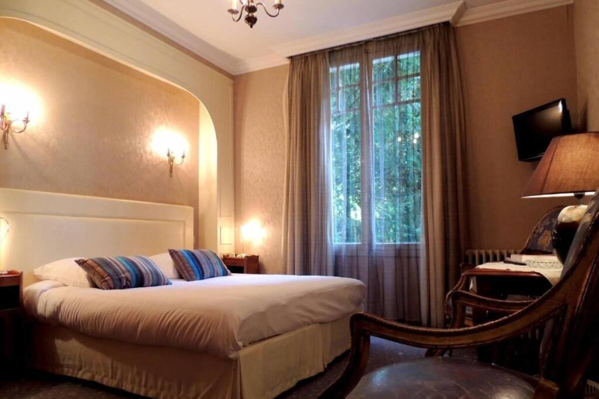 Una habitación de hotel con cama, una silla, televisor y ventana con cortinas.