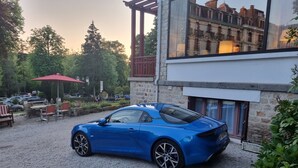 Une voiture de sport bleue est garée sur une allée de gravier, avec un bâtiment en pierre en arrière-plan.