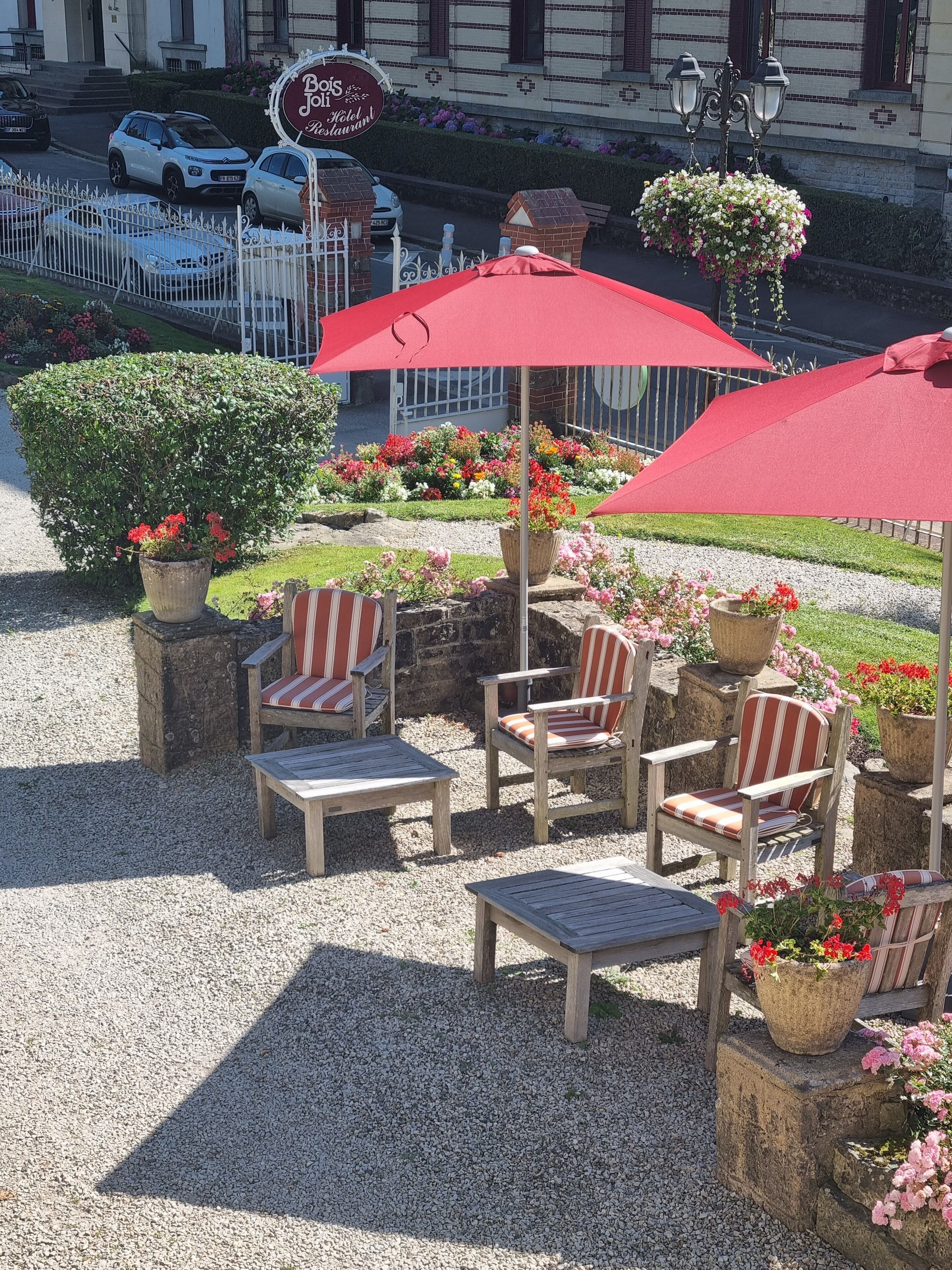 Een terras met rode parasols, stenen plantenbakken met bloemen en gestreepte stoelen.