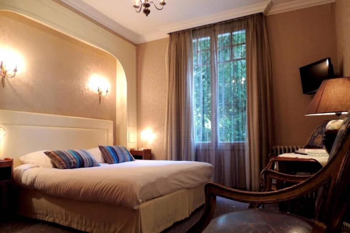 Una habitación de hotel con cama, una silla, televisor y ventana con cortinas.