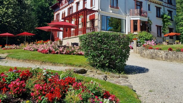 Un bâtiment traditionnel à colombages, doté d’un balcon, entouré d’un jardin bien entretenu où poussent différentes espèces de fleurs et qui est agrémenté d’un chemin de gravier.