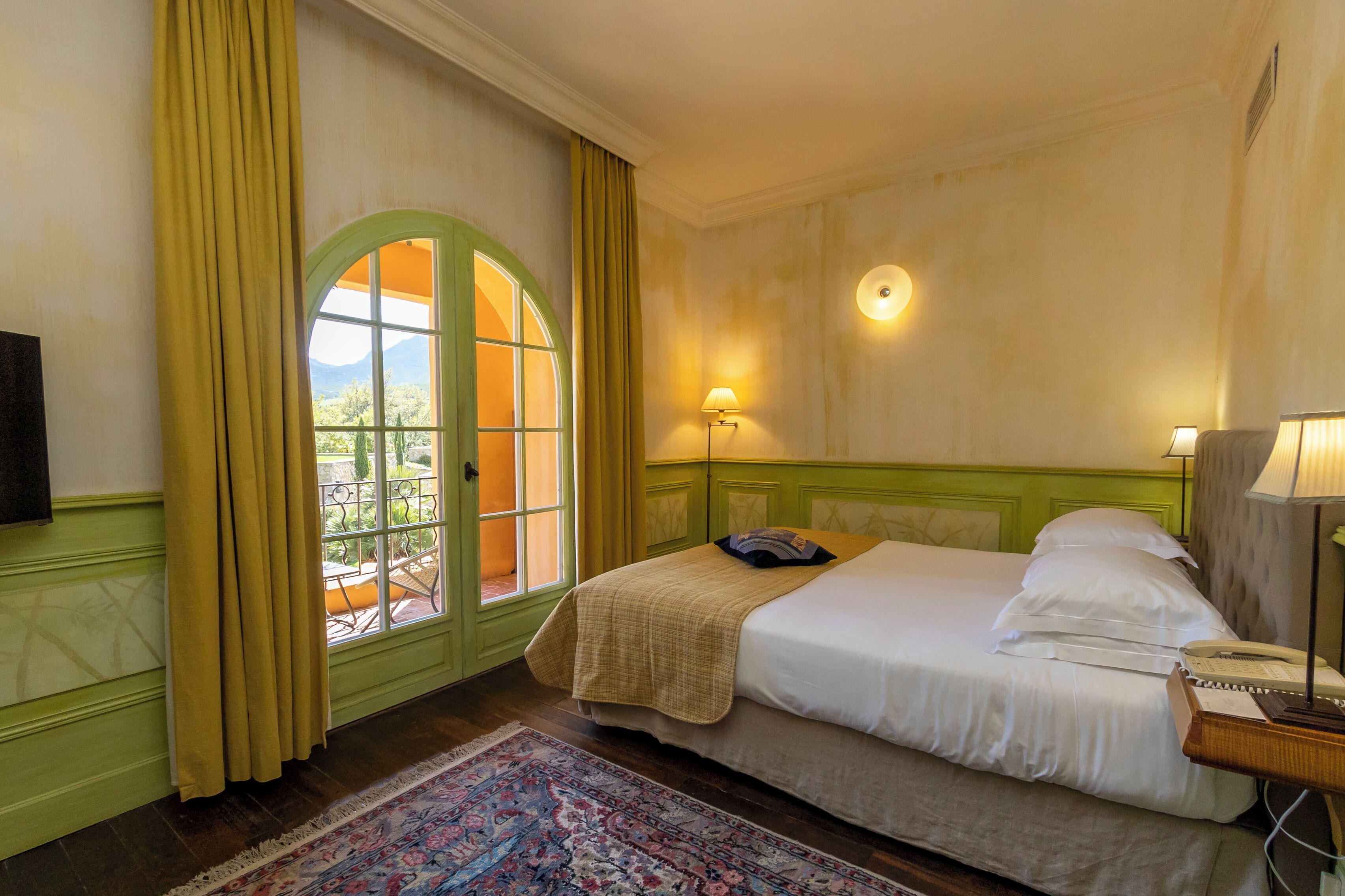 A hotel room with a bed, a nightstand, a lamp, a window with curtains, and a view of the outdoors.