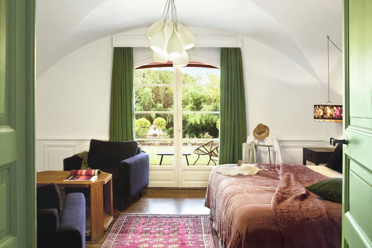Un dormitorio con un ventanal grande, una cama, una silla, una mesa y una lámpara colgante.