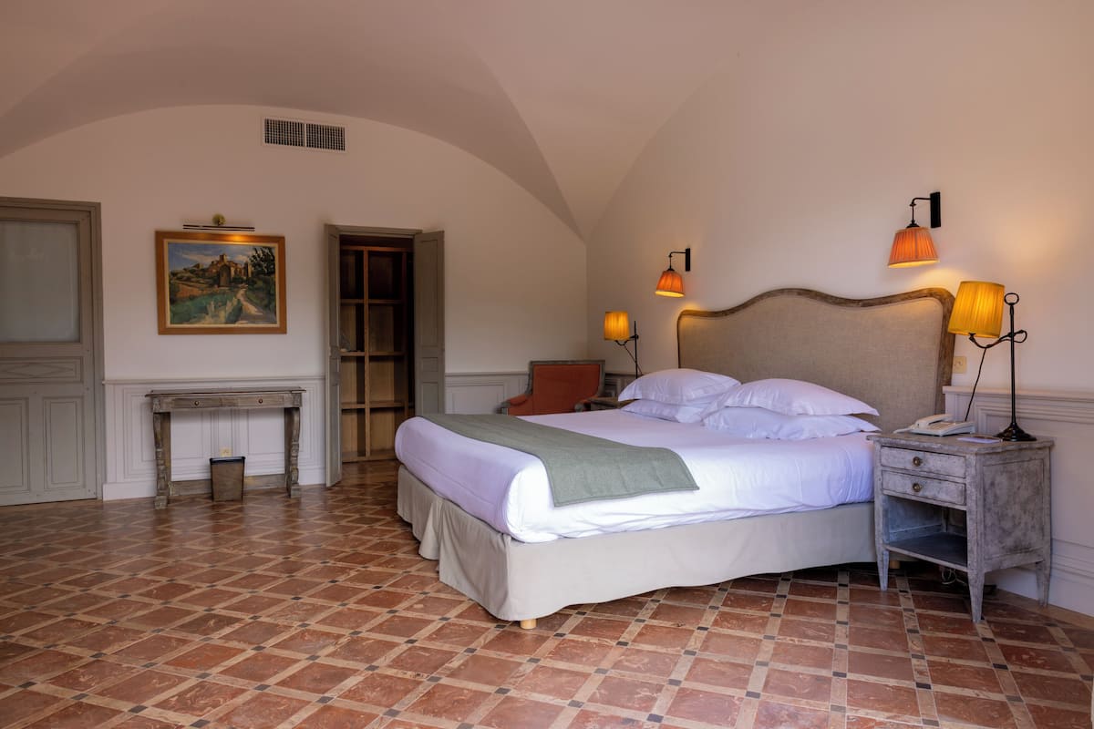 Un dormitorio con cama, dos lámparas de noche, una mesita de noche, una silla, un cuadro y una puerta.