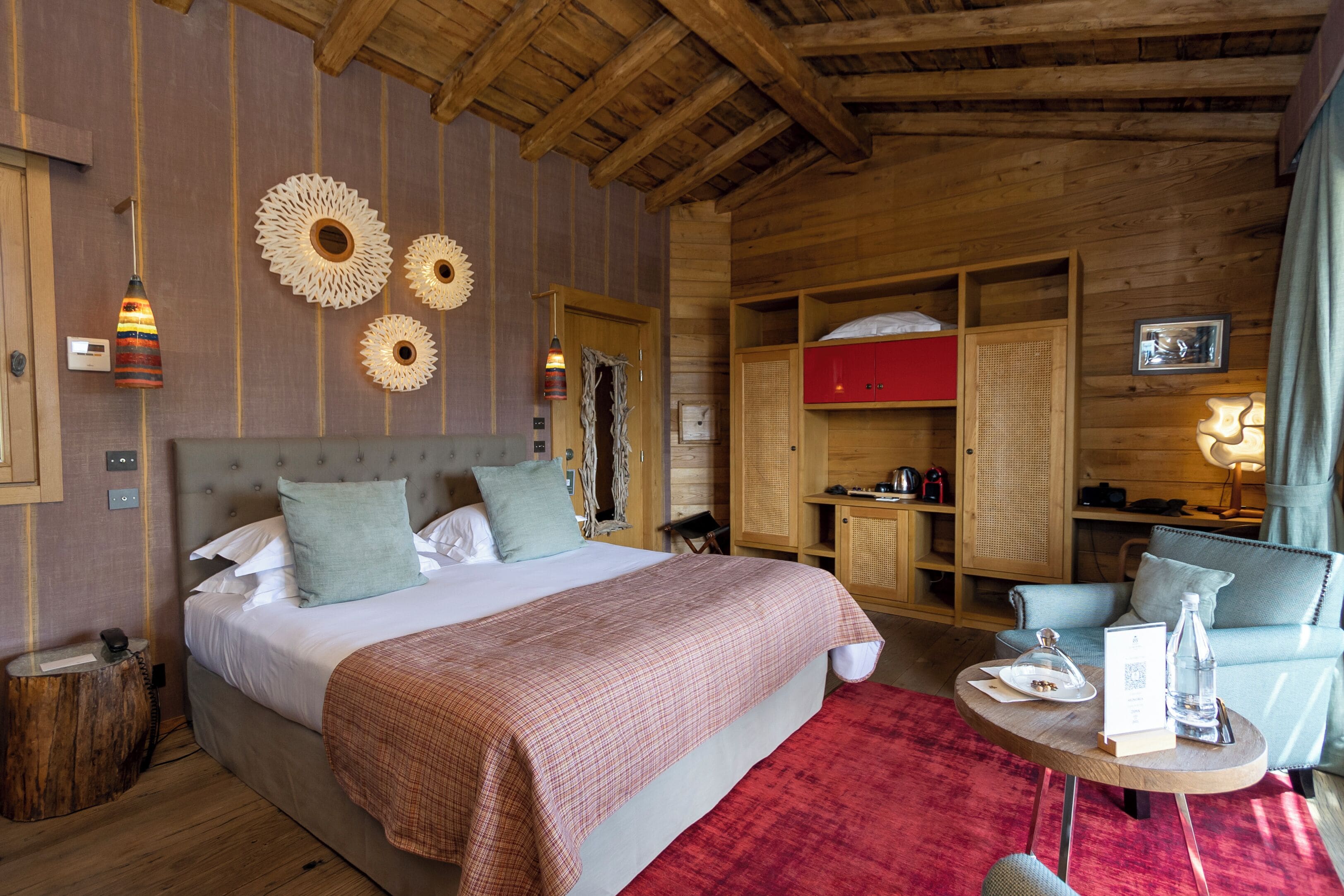 Une pièce chaleureuse avec un grand lit, un plafond en bois et une moquette rouge. On y trouve une petite table avec une bouteille et un verre, ainsi qu’une chaise avec un coussin.