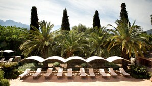 Zona de piscina con sillones de descanso y sombrillas, rodeada de palmeras y cipreses, con montañas al fondo.