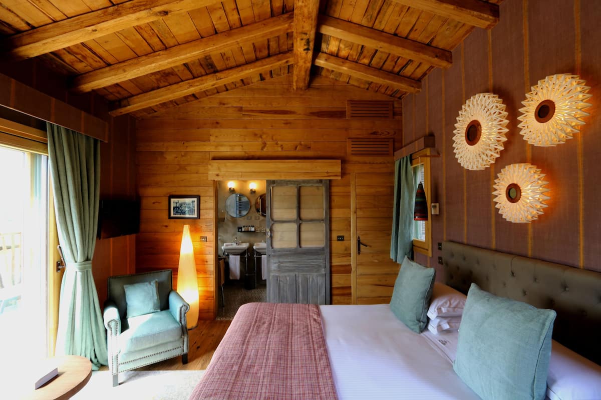 Habitación de una cabaña de madera con una cama, dos sillones, una lámpara y un baño a la vista, visible a través de una puerta abierta.