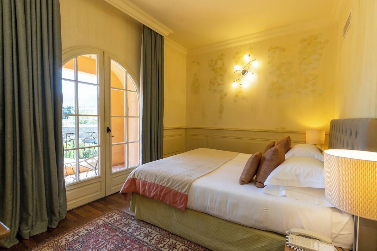 Habitación de hotel con una cama grande, una ventana con cortinas, una lámpara de pared y un tapete estampado.
