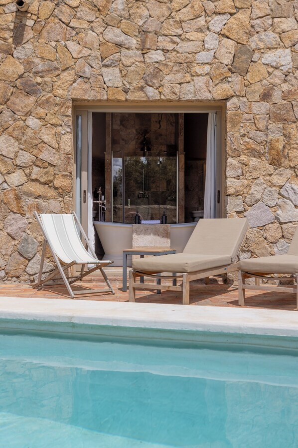 A stone wall with a glass door leading to a bathroom. There are two lounge chairs and a small table by a pool.