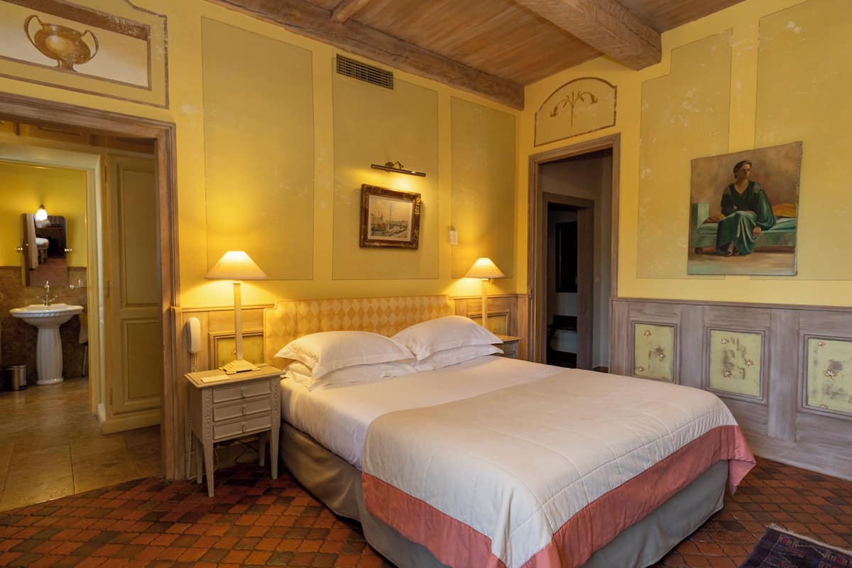 Un cuarto de hotel con una cama grande, dos lámparas de noche, una mesita de noche, un cuadro en la pared y un cuadro sobre la cama.