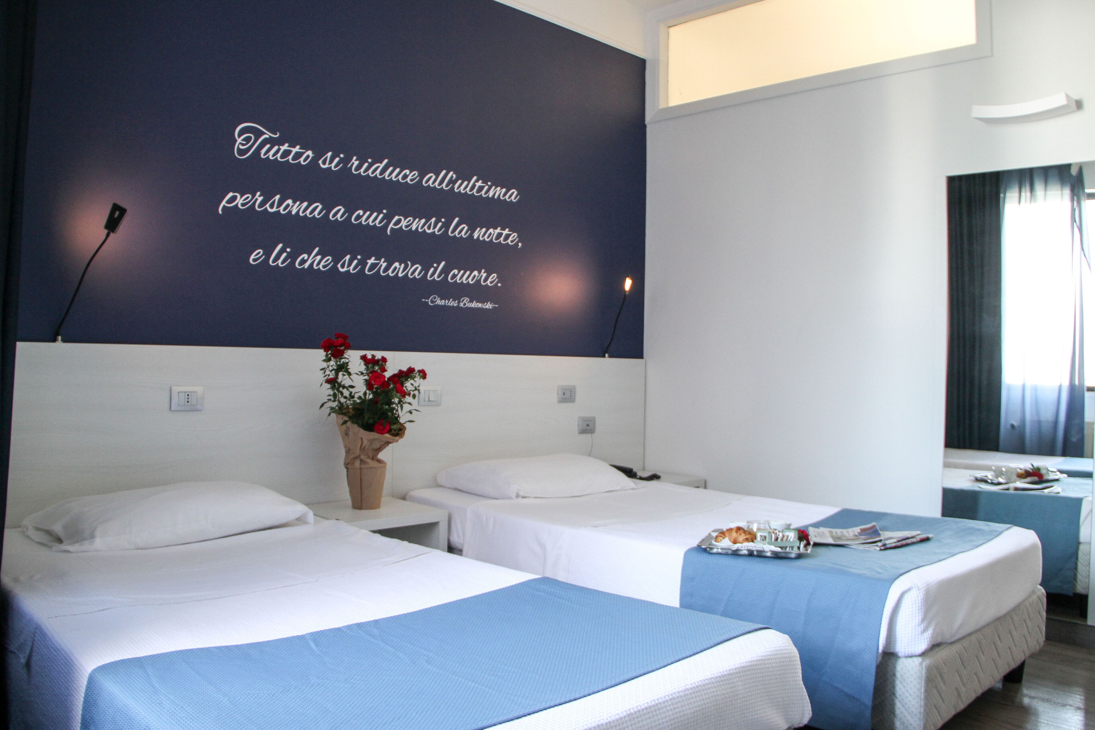 Une chambre d’hôtel avec deux lits, une grande citation affichée sur le mur, un vase avec des fleurs et un plateau de petit-déjeuner sur le lit de droite.