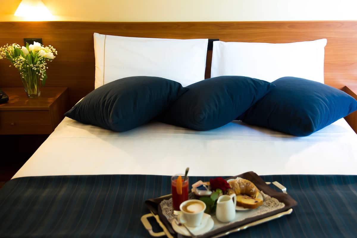 Un lit bien fait, orné d’oreillers bleu foncé, un plateau avec des articles pour le petit-déjeuner et un vase de fleurs sur la table de chevet.