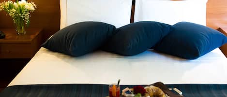Una cama bien hecha con almohadas azul oscuro, una bandeja con alimentos para el desayuno y un florero sobre la mesita de noche.