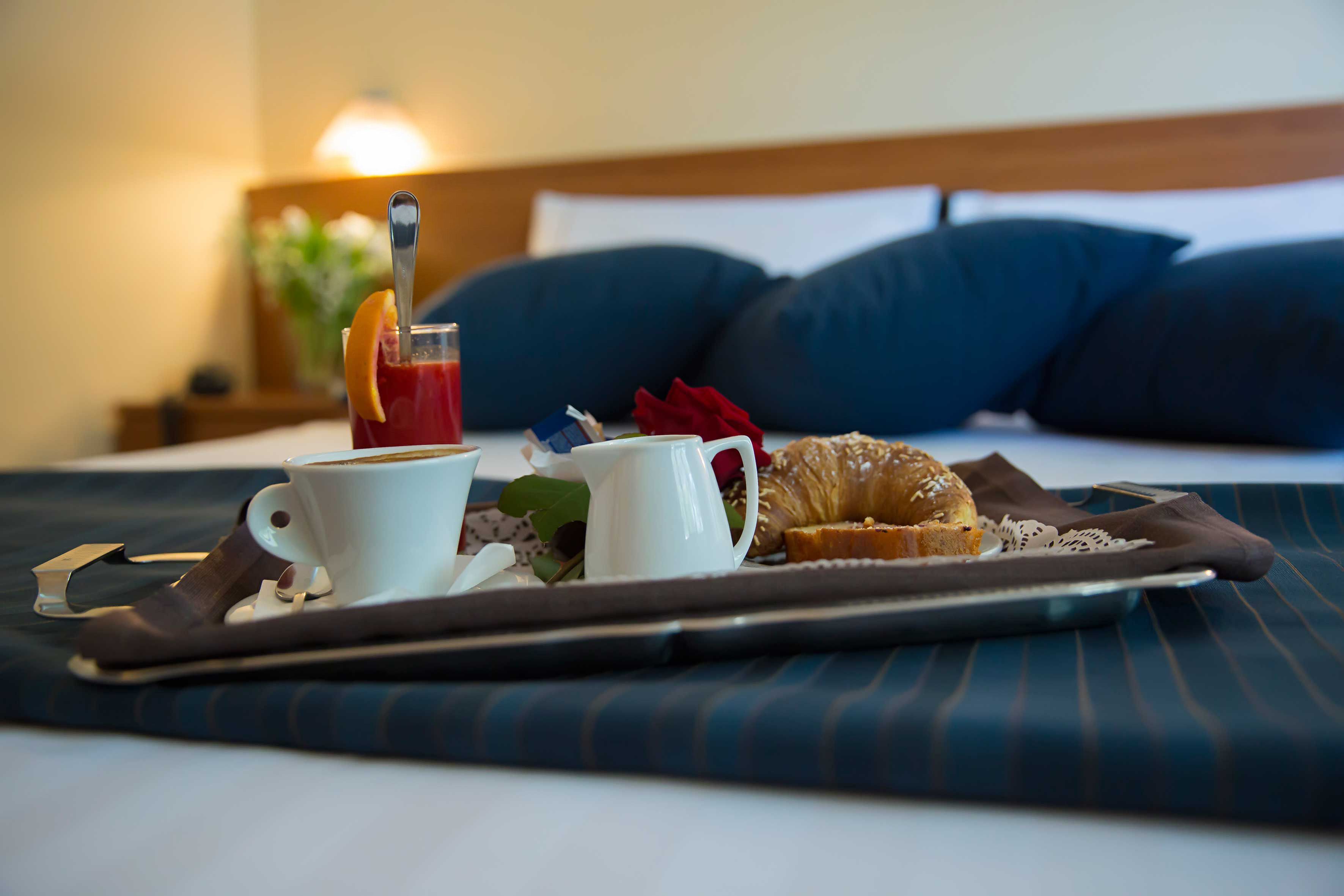 A breakfast tray with a cup of coffee, a croissant, and a glass of juice on a bed with blue bedding and a wooden headboard.