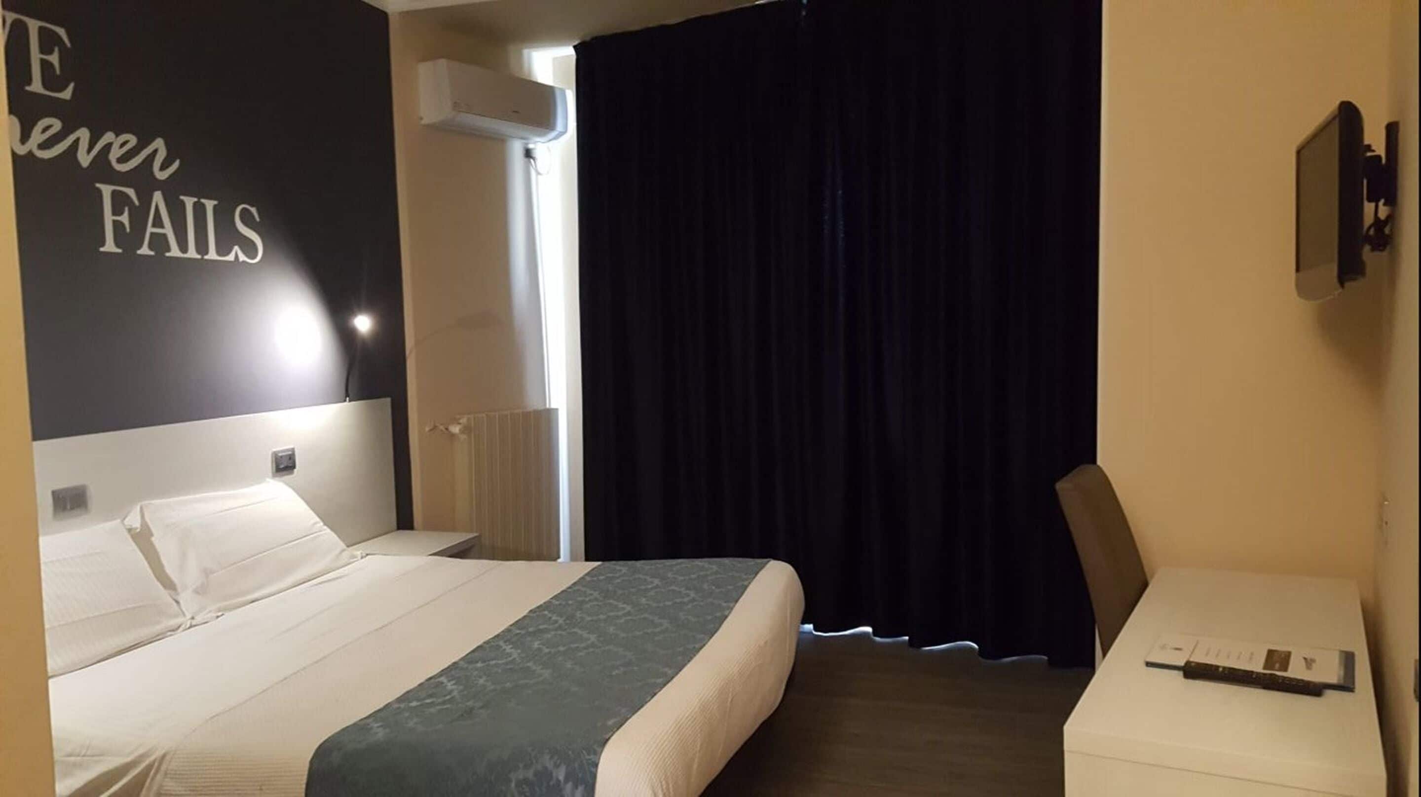 A hotel room with a bed, a desk, a chair, a black curtain, and a motivational quote on the wall.