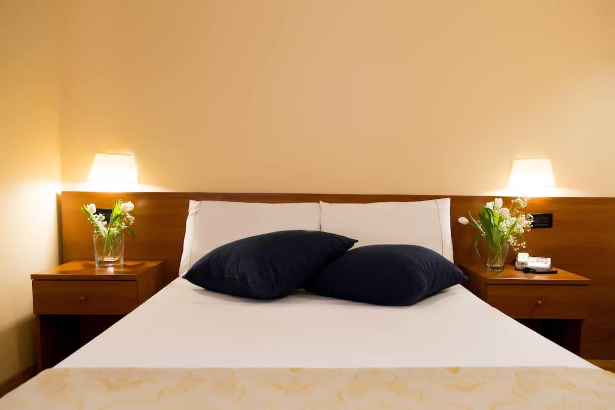 Un lit bien fait, avec deux oreillers, une table de chevet sur laquelle se trouve une lampe et un vase de fleurs, et un petit tiroir.