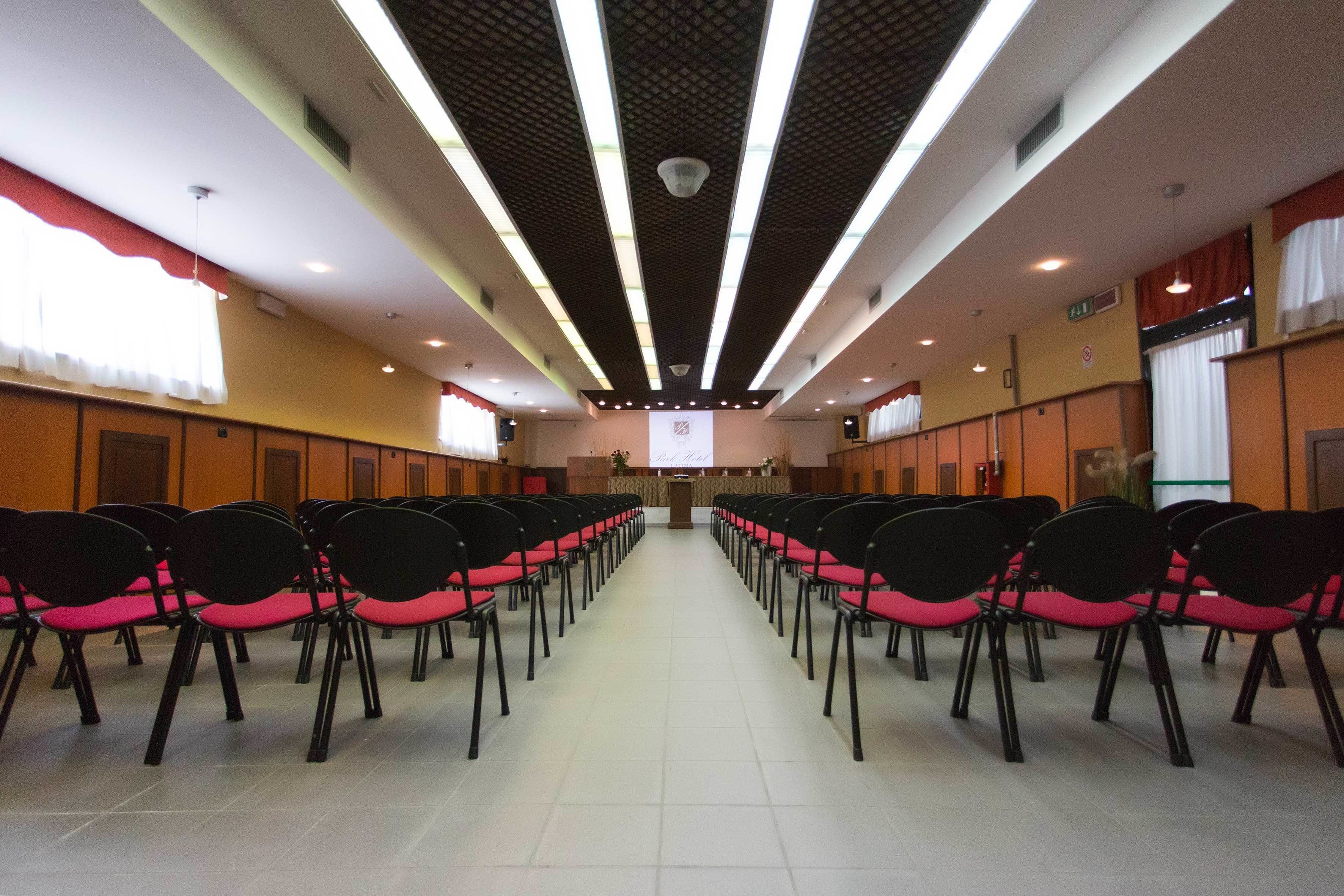 A conference room with rows of black chairs with red cushions, a podium at the front, and a projector screen.