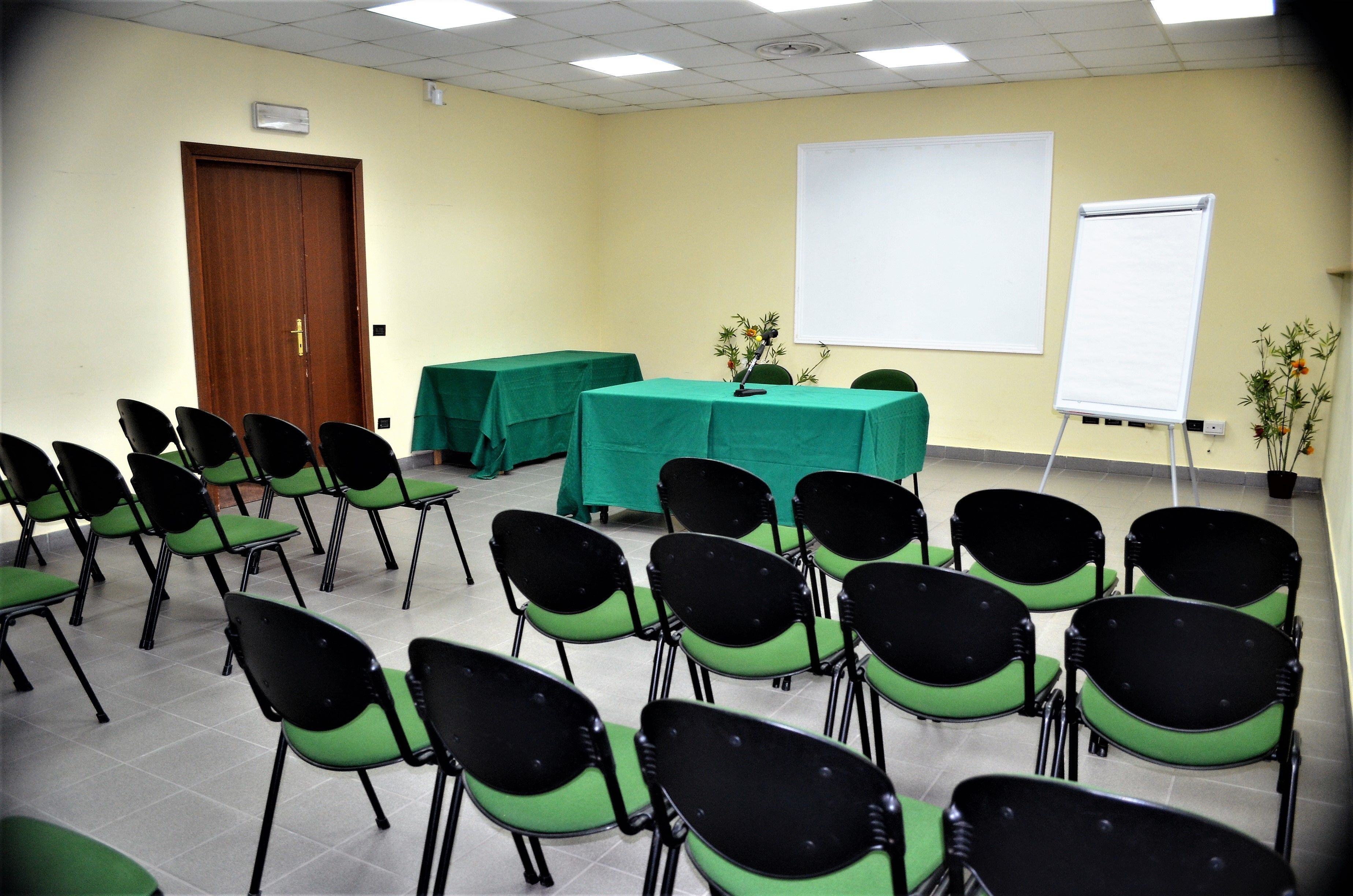 A conference room with rows of chairs, a table with a green tablecloth, and two whiteboards on the walls.