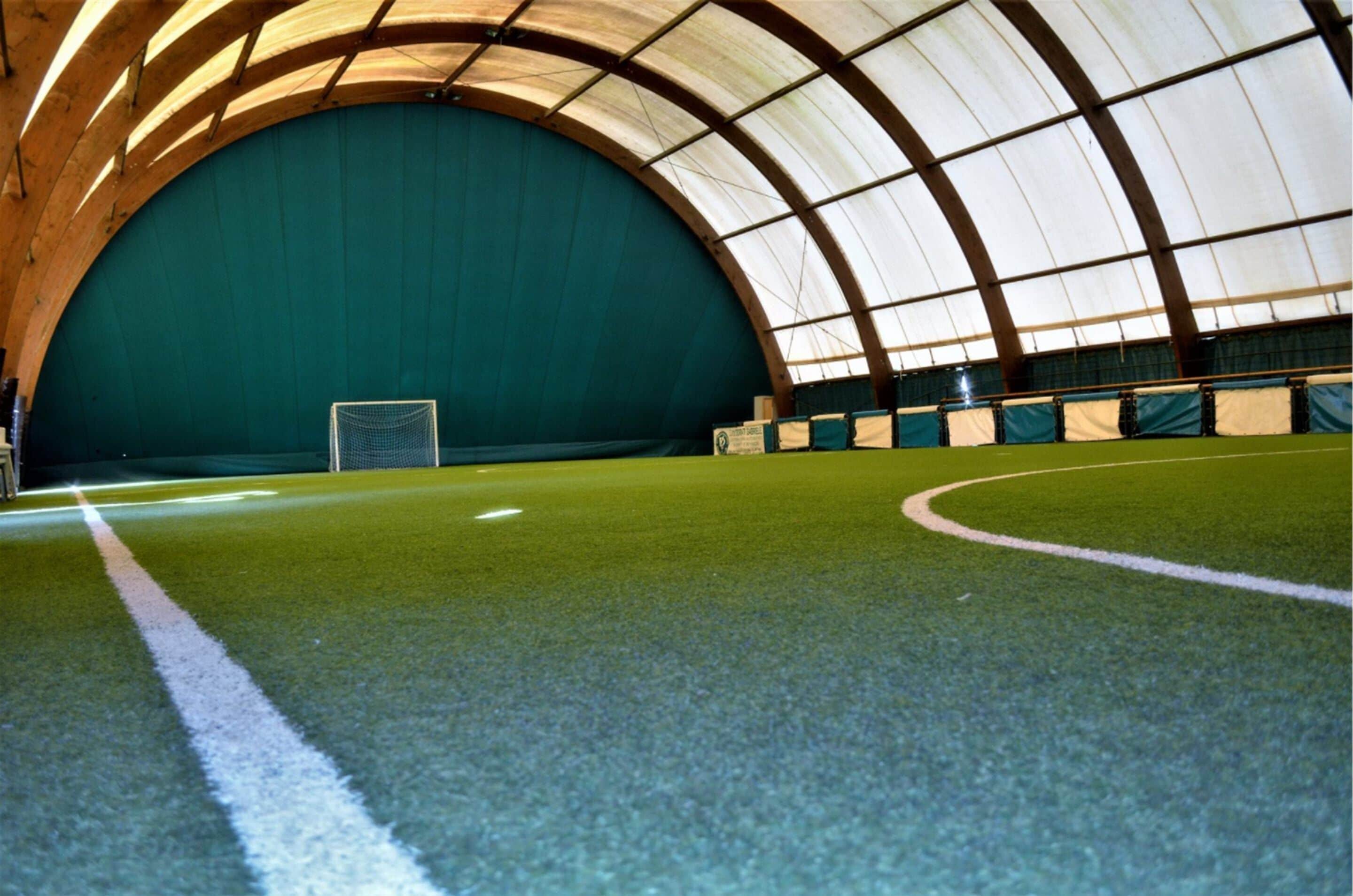 An indoor sports facility with a green turf field, goalpost, and a curved roof structure.