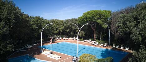 Piscina rectangular con solárium de baldosas, rodeada de árboles y equipada con múltiples sillones y tumbonas.