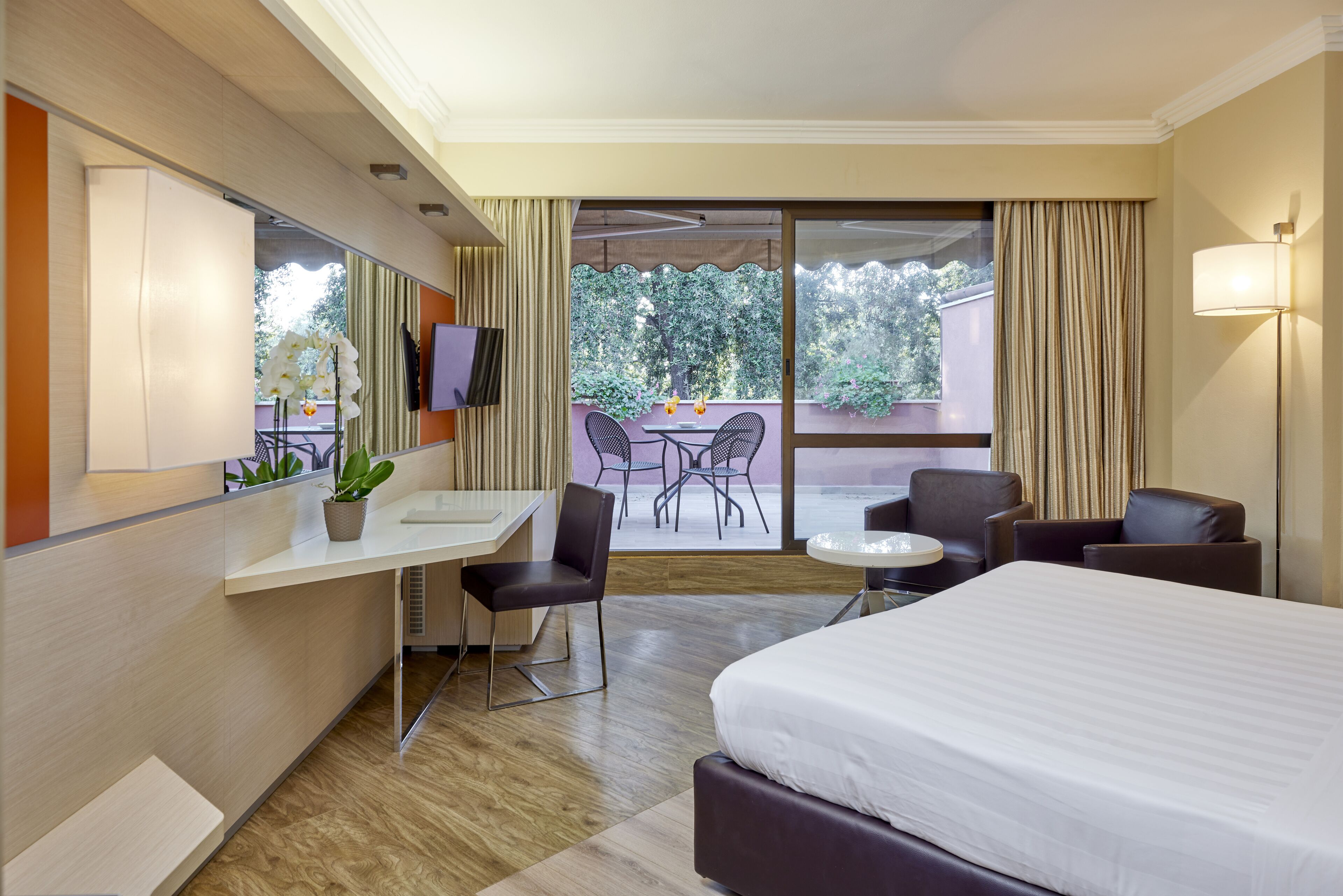 A hotel room with a bed, desk, chairs, a TV, and a balcony with a table and chairs.
