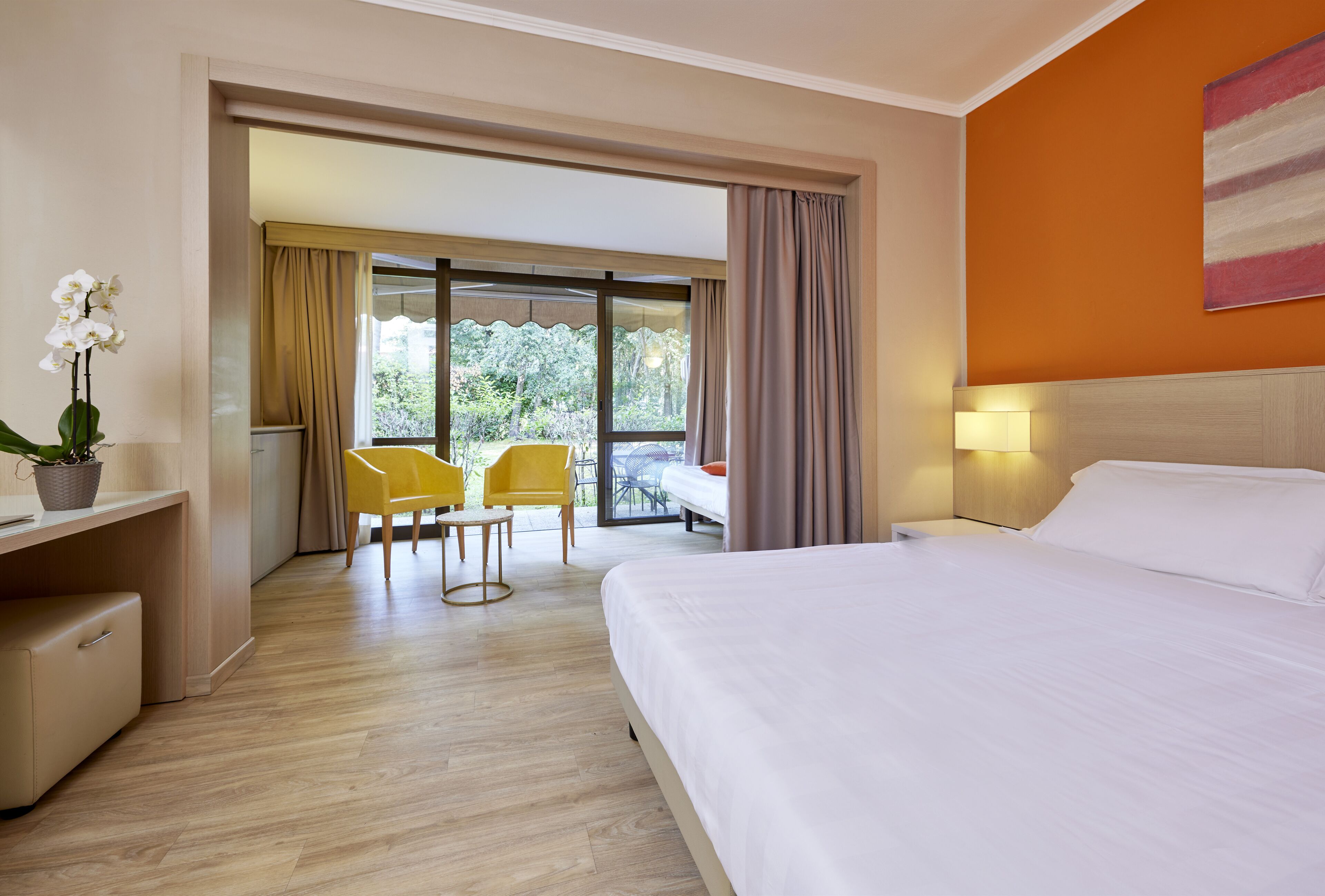 Ein Hotelzimmer mit Bett, Schreibtisch mit Pflanze, Stuhl und Blick ins Freie durch ein großes Fenster.