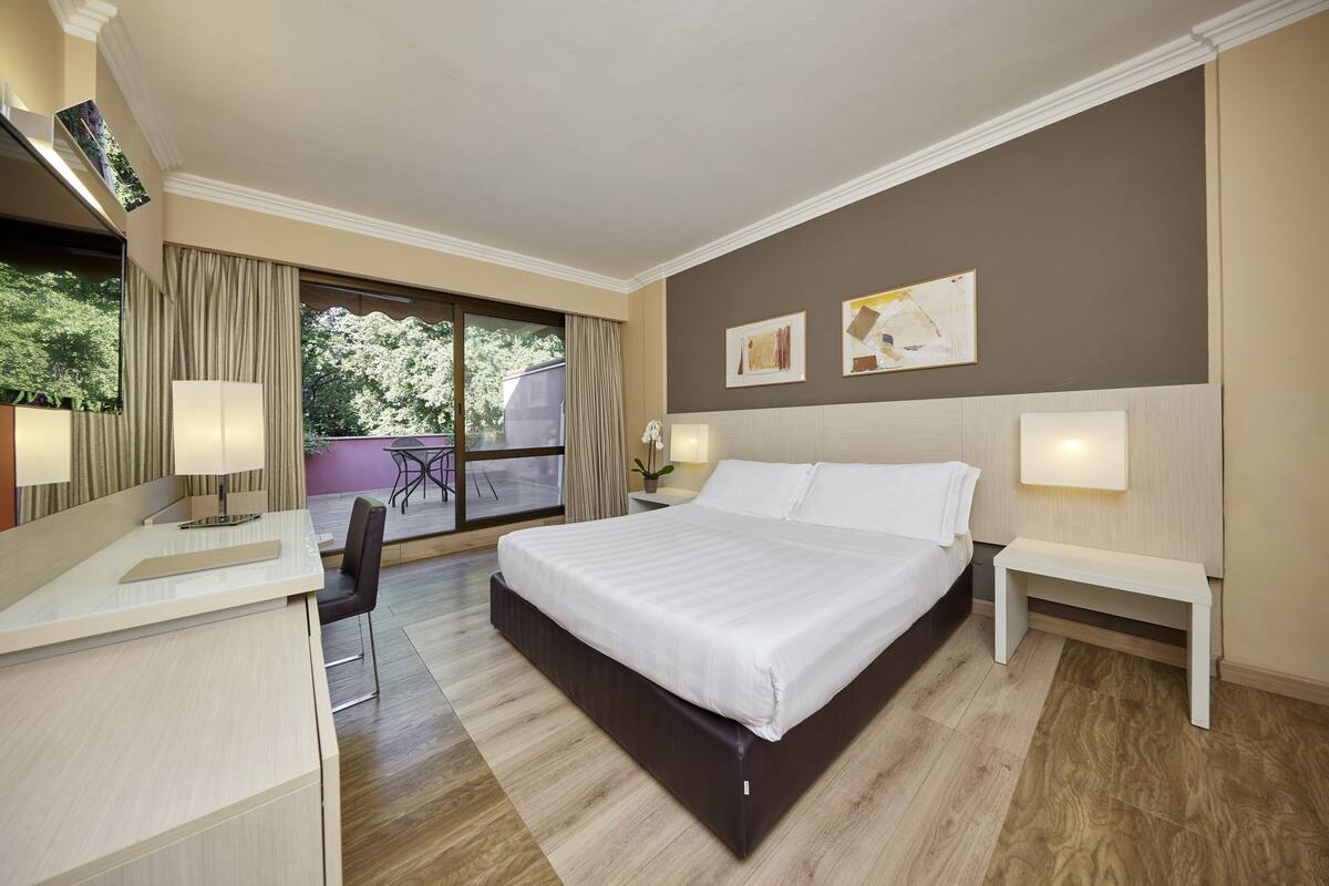 Una habitación de hotel moderna con una cama grande, mesitas de noche, un escritorio con silla y vistas al exterior.