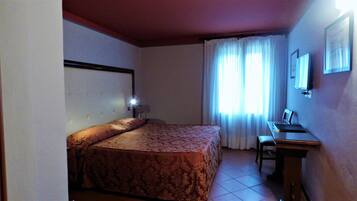 A hotel room with a bed, a desk, a chair, a lamp, and a window with curtains.