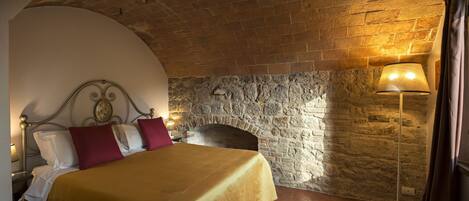 A bedroom with a stone wall, a bed with a gold bedspread, and a nightstand with a lamp.
