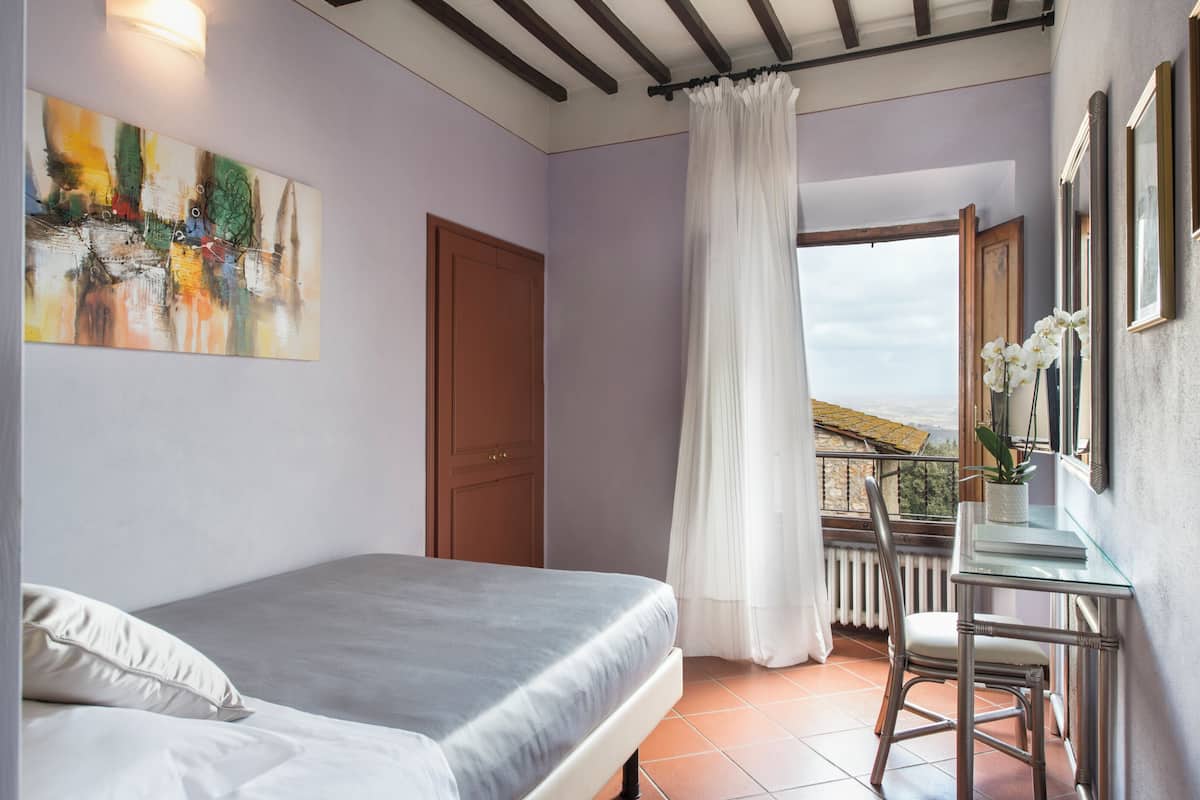 A bedroom with a bed, a desk with a chair, a window with a view, and a painting on the wall.