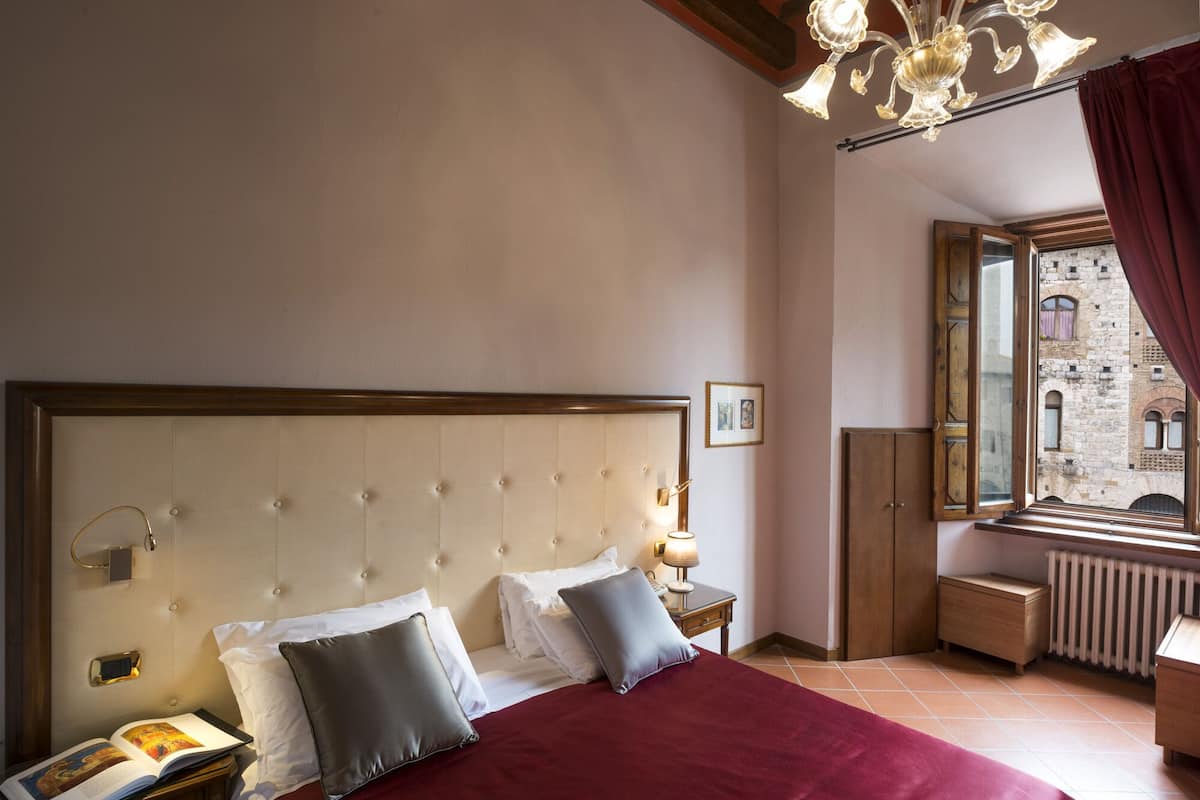 A hotel room with a large bed, a bedside table with a lamp, a window with a view of a building, and a wooden headboard.