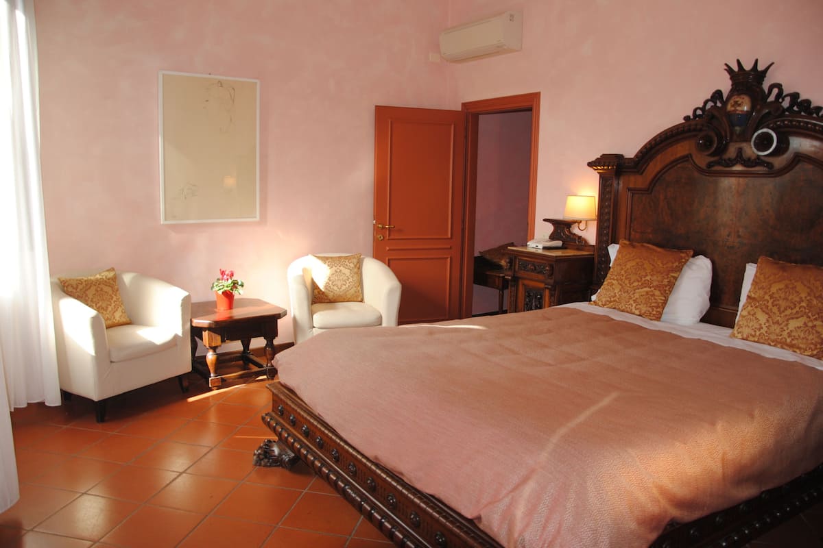 A bedroom with a large bed, two armchairs, a small table with a lamp, and a door leading to another room.