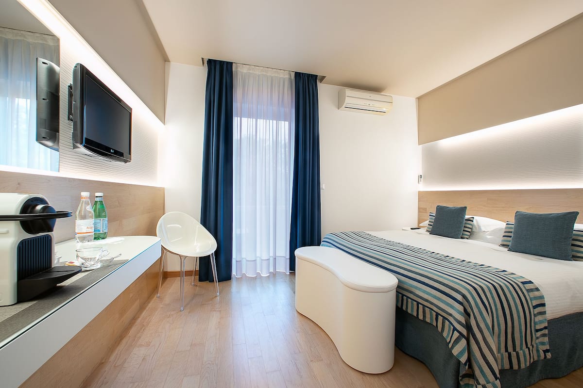 Une chambre d’hôtel moderne équipée d’un téléviseur à écran plat, d’une machine à café, d’un coin repas avec une chaise, d’un grand lit avec une couverture à rayures et d’une fenêtre avec des rideaux bleus.