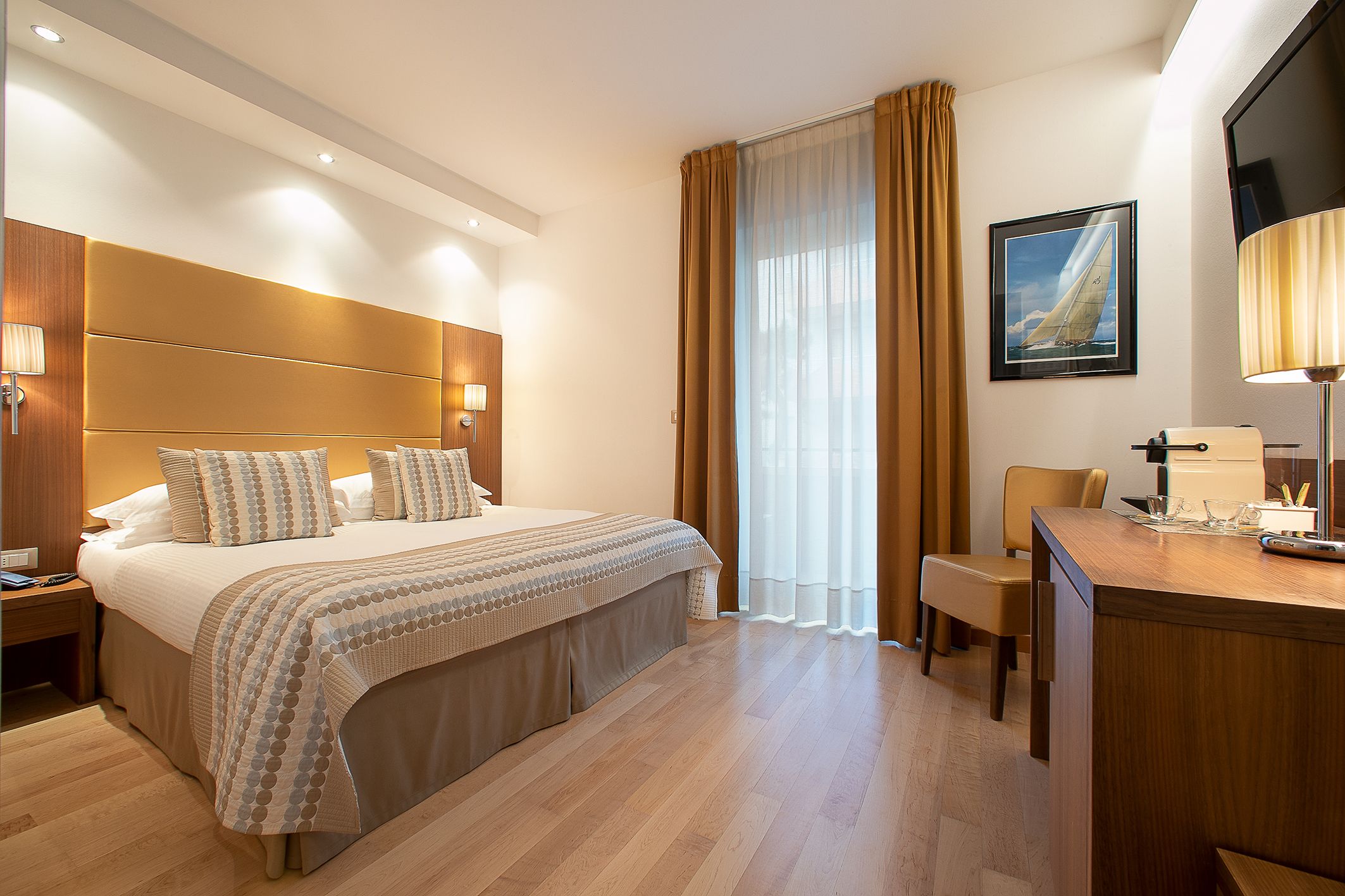 Quarto de hotel com uma cama grande, uma escrivaninha com luminária, uma cadeira e uma janela com cortinas.