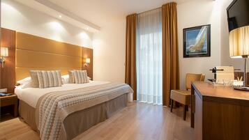 Quarto de hotel com uma cama grande, uma escrivaninha com luminária, uma cadeira e uma janela com cortinas.
