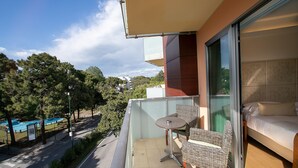 Un balcón con mesa y sillas, una cama con cabecera y vistas a una piscina y árboles.