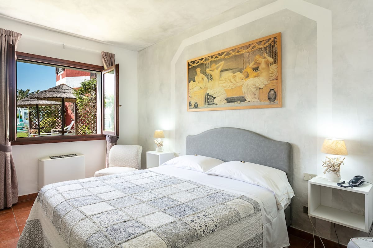 Un dormitorio con una cama grande, una mesita de noche con una lámpara, un cuadro en la pared y una ventana con cortinas.