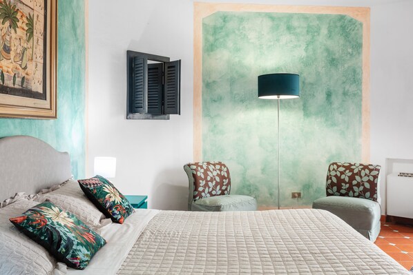 Un dormitorio con una cama, dos sillas, una lámpara de pared y un cuadro enmarco.