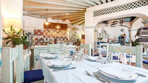 Comedor con mesas dispuestas para el servicio, cubiertos con manteles blancos, sillas tapizadas en azul y un techo decorativo con vigas a la vista.
