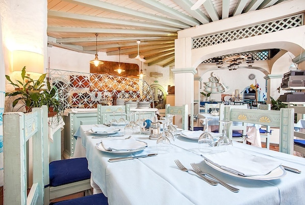A dining area with tables set for service, featuring white tablecloths, blue upholstered chairs, and a decorative ceiling with exposed beams. - Hotel Stefania Boutique Hotel by the Beach (Olbia)