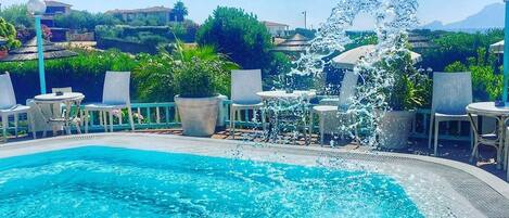 Une piscine à l’eau d’un bleu cristallin, entourée de chaises longues et de tables, avec une fontaine au centre.
