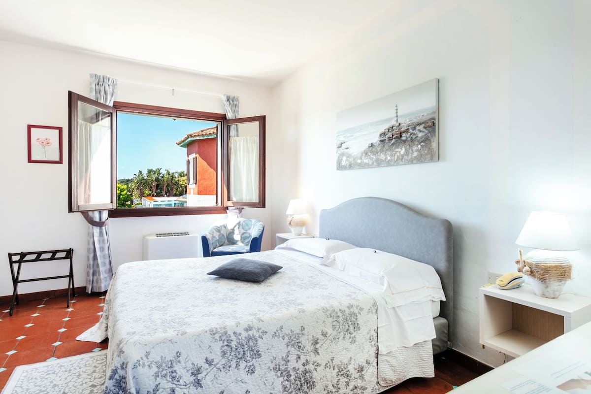 Un dormitorio con una cama grande, una ventana con cortinas, una mesita de noche con lámpara y un cuadro enmarcado en la pared.