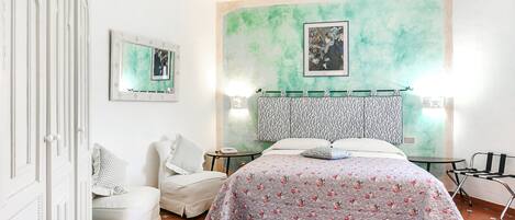A bedroom with a bed, a chair, a bedside table, and a wall with a painting.